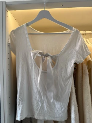 Vit topp med rosett och öppen rygg - Snygg vit topp i bomull med korta ärmar och en djup, öppen rygg som knyts med en bred satinrosett. Enkel framtill och riktigt fin detalj bak. Perfekt för dig som gillar stilrena plagg med en twist.