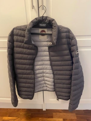 Grå pufferjacka från Colmar - Snygg grå pufferjacka från Colmar med quiltad design och hög krage. Jackan har dragkedja framtill, två sidofickor med dragkedja och Colmar-logga på ärmen. Perfekt för kalla vinterdagar och riktigt lätt att bära.