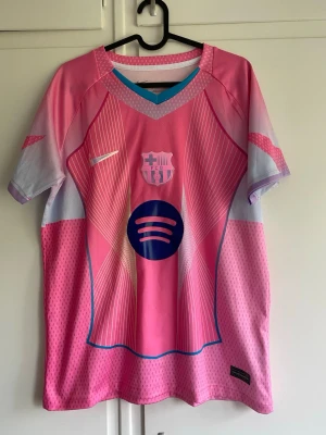 Rosa Barcelona matchtröja - Säljer en rosa och lila Barcelona matchtröja med vita och blå detaljer. Tröjan har klubbmärke och sponsorlogga på bröstet samt ett sportigt mönster framtill. Tillverkad i lätt och ventilerande funktionsmaterial, perfekt för träning eller match.