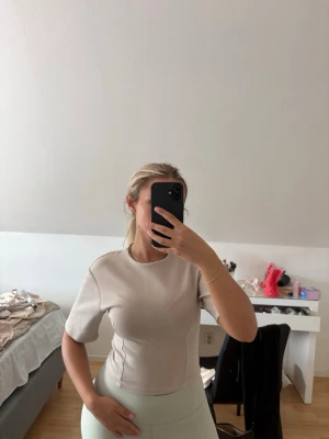 Beige tight topp med kort ärm - Säljer en stilren beige topp med tight passform och korta ärmar. Toppen har rund halsringning och markerade sömmar längs sidorna för en snygg siluett. Perfekt att matcha med både jeans och kjol. Knappt använd. Nypris 260kr.