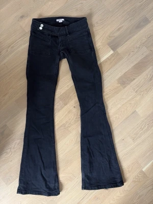 Svarta bootcut jeans med detaljer - Säljer ett par svarta bootcut jeans med två knappar i midjan och snygga fickor fram. På bakfickan finns en broderad detalj med svarta nitar som ger extra edge. Jeansen har utsvängda ben och är gjorda i ett mjukt denimtyg.