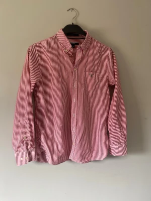 Rödvit randig skjorta från Gant - Klassisk rödvit randig skjorta från Gant med knappkrage och bröstficka med broderad logga. Skjortan har långa ärmar och vita knappar. Perfekt för en stilren och avslappnad look. Pris går att diskuteras vid snabb affär 