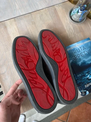 Gråa Louboutin sneakers med röd sula - Snygga sneakers från Christian Louboutin i grå mocka med den ikoniska röda sulan och logotypen undertill. Skorna har klassisk låg siluett och neutrala snören. Insidan är beige och märkt med guldfärgad logga.