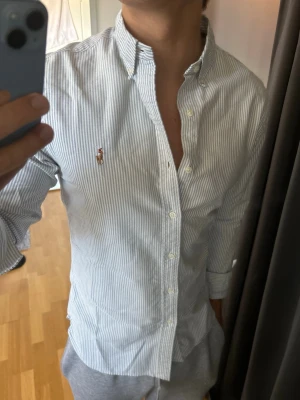 Randig skjorta med knappar - Snygg vit skjorta med tunna blå ränder och klassisk button-down krage. Skjortan har långa ärmar och knäppning framtill samt en liten broderad logga på bröstet. Perfekt för en clean och stilren look.