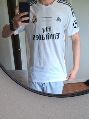 Real Madrid matchtröja Sergio Ramos - Säljer en vit Real Madrid matchtröja från Adidas med Sergio Ramos och nummer 4 på ryggen. Tröjan har svarta detaljer, Champions League-märke på ärmen och sponsortryck på bröstet. Perfekt för fotbollsfans som vill visa sitt stöd för laget.