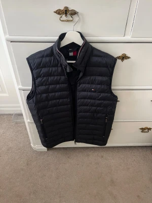 Marinblå dunväst från Tommy Hilfiger - Snygg dunväst från Tommy Hilfiger med ståkrage och dragkedja framtill. Västen har två fickor med dragkedja och diskret logga på bröstet. Perfekt för lager på lager och ger en sportig look.