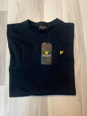 Svart t-shirt från Lyle & Scott - En stilren svart t-shirt från Lyle & Scott med klassisk rund hals och korta ärmar. T-shirten har den ikoniska gula logon broderad på bröstet och är tillverkad i mjuk bomull för skön komfort. Perfekt basplagg för en clean look.