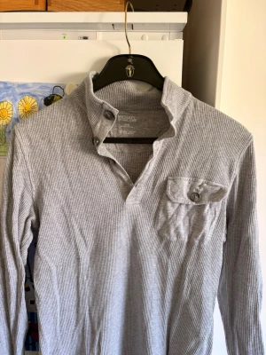 Grå långärmad tröja Michael Kors - Snygg grå långärmad tröja från Michael Kors i ribbad bomull. Tröjan har en unik krage med knappar och en bröstficka med lock och knapp. Perfekt för dig som gillar stilrena och bekväma plagg.