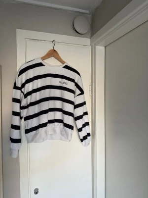 Randig sweatshirt med BALANCE-tryck - Säljer en vit sweatshirt med svarta horisontella ränder och BALANCE-tryck på bröstet. Tröjan har rund halsringning, långa ärmar och ribbade muddar vid ärmslut och nederkant. 