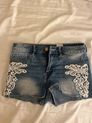 Blå jeansshorts med spetsdetaljer - Säljer ett par blå jeansshorts med rå kant och vita spetsapplikationer på framsidan. Shortsen har fem fickor, knapp och dragkedja framtill samt en klassisk denimlook med slitningar. Perfekta för sommaren och ger en cool, avslappnad vibe.