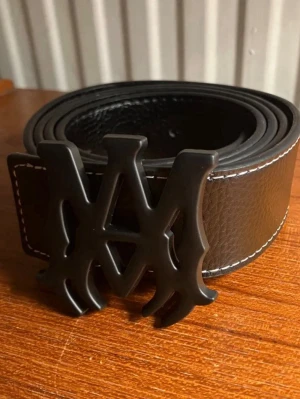 Svart skärp AMIRI - Snyggt svart skärp från AMIRI iskinn med ett stort, svart metallspänne i form av bokstäver. Skärpet har vita kontrastsömmar längs kanterna och en stilren, modern look som passar till jeans eller byxor. Helt nytt, kommer med hålgörare 