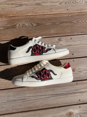 Vita sneakers med orm och ränder - Säljer ett par vita sneakers med coolt ormmotiv på sidan och klassiska gröna och röda ränder. Skorna har vita snören, låg profil och detaljer i rött och grönt på hälen. Tillverkade i slätt läder för en clean look.