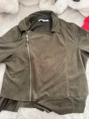 Olivgrön skinnjacka med dragkedjor - Snygg olivgrön skinnjacka med asymmetrisk dragkedja framtill och dragkedjor vid ärmsluten. Jackan har en modern krage och raka linjer som ger en cool look. Perfekt för dig som vill ha en stilren och trendig jacka till din outfit.