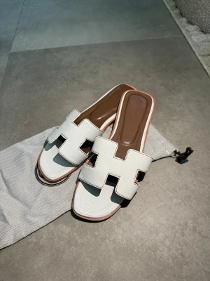 Vita slip-in sandaler med bred rem - Snygga vita sandaler med breda remmar i läder och öppen tå. Modellen har en fyrkantig tå och bruna innersulor för extra kontrast. Perfekta för sommaren och lätta att matcha till olika outfits. Klassisk design med trendig touch.