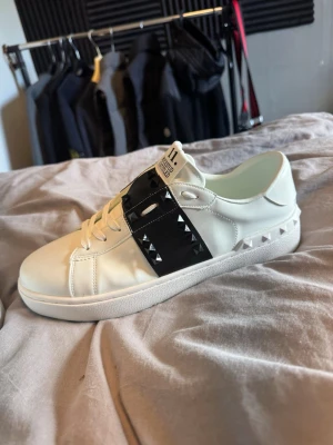 Vita Valentino Garavani sneakers - Säljer ett par vita Valentino Garavani rockstuds sneakers med svart band och silverfärgade nitar på sidan. Skorna har en klassisk låg profil, vita snören och är tillverkade i slätt läder. Perfekta för dig som vill ha en stilren men edgy look.
