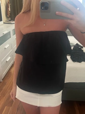 Svart axelbandslös tubtopp - Snygg svart axelbandslös tubtopp med volangdetalj upptill. Perfekt till sommaren och enkel att matcha med olika outfits. Toppens material känns mjukt och lätt mot huden.