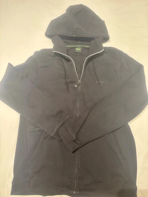 Svart hoodie med dragkedja - Säljer en svart hoodie med dragkedja och huva. Tröjan har långa ärmar, dragsko i huvan och fickor på sidorna. Perfekt för en avslappnad och stilren look. Materialet känns mjukt och bekvämt, och passformen är klassisk.