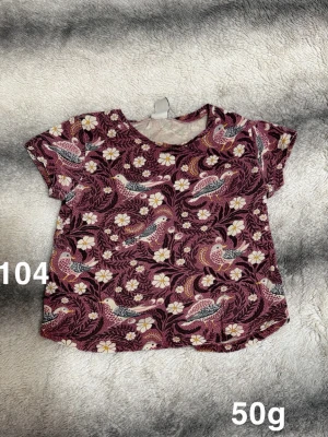 Vinröd t-shirt med fågelmönster - Kortärmad t-shirt i vinrött med ett detaljerat mönster av fåglar och vita blommor. Rund halsringning. Perfekt för dig som gillar färgglada och lekfulla prints.🌸 Fint använt skick. Storlek 104