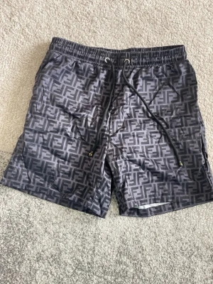 Fendi shorts- bad shorts- style - Säljer ett par svarta shorts med grått grafiskt mönster och snörning i midjan. Shortsen har elastisk midja och detaljer i metall på snöret. Perfekta för sommaren och har en loose passform.