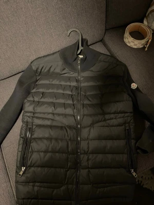 Svart pufferjacka från Moncler - Snygg svart pufferjacka från Moncler med quiltad front och stickade ärmar. Jackan har två dragkedjeförsedda fickor och en Moncler-logga på ärmen. Perfekt för kalla vinterdagar och ger en clean, modern look.
