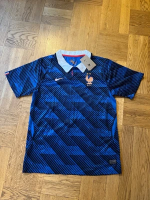 Blå Nike Frankrike fotbollströja - Säljer en blå fotbollströja från Nike med Frankrikes landslagsmärke och två stjärnor på bröstet. Tröjan har korta ärmar, vit krage och ett diagonalrandigt mönster i olika blå nyanser. Perfekt för dig som älskar fotboll och vill stötta Les Bleus!