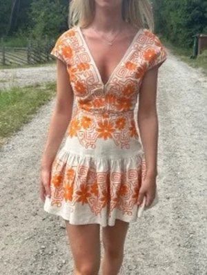 Kort klänning med orange blommönster - Säljer en kort klänning i beige med stora orange blommor och grafiskt mönster. Klänningen har v-ringning, korta ärmar och volang i nederkanten. Perfekt för sommarens alla tillfällen och ger en färgstark look.