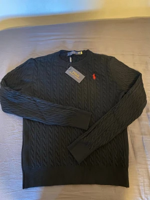 Ralph Lauren stickad tröja  - Kabelstickad svart tröja från Polo Ralph Lauren med rund halsringning och röd broderad logga på bröstet. Tröjan har långa ärmar och ribbade muddar vid ärmslut och nederkant. Klassisk och stilren design i mjukt material. Storlek S