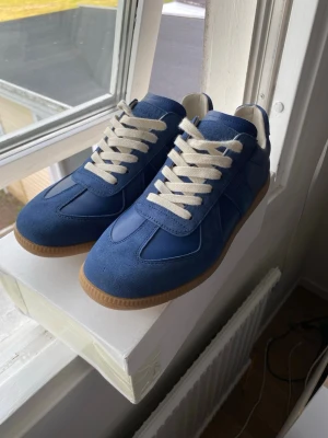 Blå sneakers med vita snören gats - Snygga blå sneakers med ovandel i mocka och släta partier, samt vita snören och beige sula. Skorna har en klassisk låg siluett och stilren design som passar till det mesta. Perfekta för dig som vill ha en clean och modern look. Snygga skor 10/10 helt nya