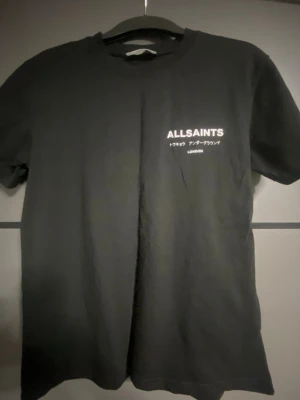 Svart AllSaints t-shirt med tryck - Säljer en svart t-shirt från AllSaints med vitt tryck både fram och bak. T-shirten har klassisk passform, rund hals och korta ärmar. Materialet är mjuk bomull som känns skön mot huden. Perfekt för en clean och stilren look.