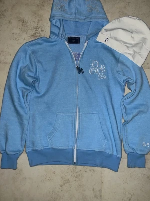 Ljusblå zip hoodie med tryck - Ljusblå hoodie med dragkedja och huva, samt vita tryckdetaljer på bröstet och ärmen där det står 'Dreese'. Mjuk bomullsmix, ribbade muddar och fickor framtill. Perfekt för en avslappnad streetwear-look.