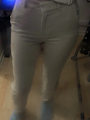 Beige rutiga kostymbyxor - Snygga beige kostymbyxor med diskret rutmönster. Byxorna har raka ben, hällor för skärp och klassisk dragkedja framtill. Materialet ser ut att vara en mjuk och lite stretchig blandning, perfekt för en clean och stilren look.