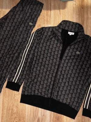 Svart mönstrad loungewear från Lacoste - Snyggt set från Lacoste i svart med grått geometriskt mönster. Jackan har dragkedja, ståkrage och vita ränder längs ärmarna. Byxorna matchar med samma mönster och har resår i midjan. Perfekt för chill dagar hemma eller när du vill vara bekväm men ändå se fräsch ut.