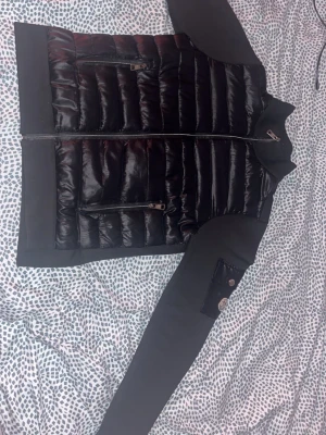 Svart pufferjacka med dragkedjor - Snygg svart pufferjacka med glansig vadderad front och två dragkedjefickor. Ärmarna är i ett mjukare tyg och har en ficka med knappdetalj på ena ärmen. Jackan har hög krage och stängs med dragkedja framtill. Inga skador på jackan kommer med tagsen  