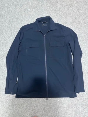 Mörkblå overshirt från Riley - Stilren mörkblå overshirt från Riley med två stora bröstfickor och dragkedja framtill. Overshirten har klassisk krage och raka ärmar med knapp vid ärmslut. Perfekt lager på lager-plagg i ett mjukt och tåligt material.