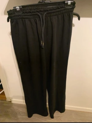 Svarta mjukisbyxor med dragsko - Säljer ett par svarta mjukisbyxor med resår i midjan och justerbar dragsko. Byxorna har raka ben och är tillverkade i ett mjukt och bekvämt material, perfekta för chill dagar. Passar dig som gillar en avslappnad stil.