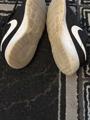 Svarta Nike sneakers med vit sula - Snygga svarta Nike sneakers med vit sula och vit swoosh på sidan. Ovandelen är i meshmaterial som andas och skorna har klassisk snörning. Perfekta för en sportig och avslappnad stil.