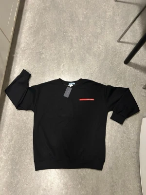 Prada, Svart sweatshirt med röd detalj - En svart Prada sweatshirt med rund halsringning och en diskret röd detalj på bröstet. Tröjan har en klassisk passform med ribbade muddar vid ärmslut och nederkant. Perfekt för en avslappnad och stilren look.