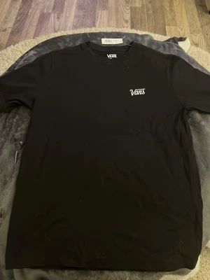 Svart Vans t-shirt med logga - Säljer en svart t-shirt från Vans med klassisk vit Vans-logga på bröstet. T-shirten har rund hals och korta ärmar, perfekt för en avslappnad och stilren look. Tillverkad i mjuk bomull för skön komfort.