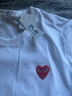 Comme des Garçons t-shirt  - Vit t-shirt från Comme des Garçons PLAY med det ikoniska röda hjärtat med ögon broderat på bröstet. Klassisk rund hals och korta ärmar. Tillverkad i mjuk bomull för en skön känsla. Perfekt för en clean och stilren look. Storlek S. Säljer eftersom köpte fel storlek 
