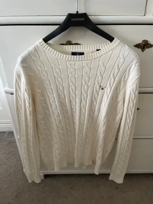 Vit kabelstickad tröja - Kabelstickad tröja i krämvit färg med rund halsringning och långa ärmar. Tröjan har ett diskret broderat emblem på bröstet och ribbade muddar vid ärmslut och nederkant. Perfekt för lager på lager och klassisk stil.