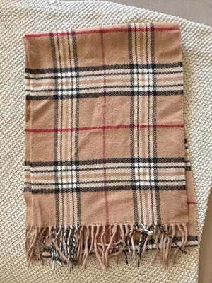 Beige halsduk med rutigt mönster - Snygg beige halsduk med klassiskt rutigt mönster i svart, vitt och rött. Halsduken har fransar i ändarna och är tillverkad i ett mjukt tyg som känns skönt mot huden. Perfekt accessoar för att lyfta din outfit.
