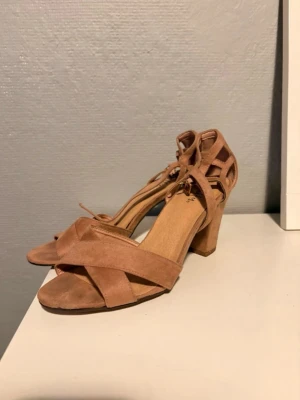 Beiga sandaler med klack och snörning - Snygga sandaler i beige mockaimitation med breda korslagda remmar framtill och snörning runt ankeln. Klackarna är stabila och mellanhöga, vilket ger både stil och komfort. Perfekta för att lyfta vilken outfit som helst.