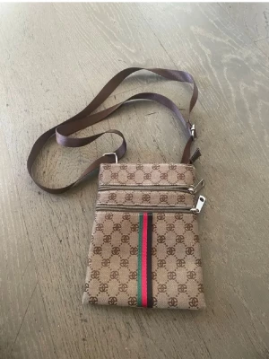 Beige axelväska med monogram och ränder - Snygg beige axelväska med brunt monogrammönster och två dragkedjefack framtill. Väskan har en brun justerbar axelrem och en dekorativ grön och röd rand på framsidan. Perfekt för att bära det viktigaste och ger en trendig look.