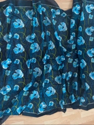 Ny. Blommig blå scarf i tunn bomull  - Snygg sjal med blommigt mönster i olika blå toner på mörk botten. Sjalen är tunn och luftig, perfekt att slänga över axlarna för en cool touch till din outfit. Blommorna har gröna stjälkar och ger ett fräscht intryck. Storlek 172x113cm. Bomull 100%
