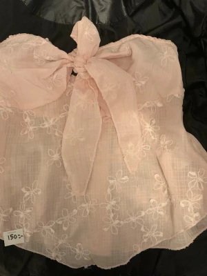 Ljusrosa axelbandslös topp med rosett xs/s  - Supersöt ljusrosa axelbandslös topp med stor rosett framtill och broderade blommor. Toppen är bandeau-modell och har smock baktill för extra komfort. Perfekt för sommarens alla tillfällen! Vet inte riktigt vart den är ifrån då lappen är avklippt men skulle gissa bik bok. Är tunt material och kan vara i bomull. 