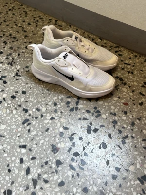 Vita Nike sneakers med svart swoosh - Fräscha vita Nike sneakers med svart swoosh på sidan. Skorna har en sportig och clean design med mesh- och syntetmaterial för bra andningsförmåga. Lågt skaft och tjock sula för skön komfort. Perfekta för dig som gillar stilrena och klassiska sneakers. Sko snören får du med men de får du lägga själv på skorna. 