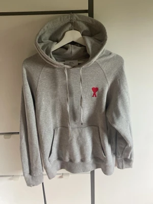 Grå Ami Paris hoodie - Snygg grå hoodie med huva och dragsko. På bröstet finns ett broderat rött hjärta med bokstaven A. Hoodien har känguruficka framtill och ribbade muddar vid ärmar och nederkant. Perfekt för en chill och avslappnad stil.