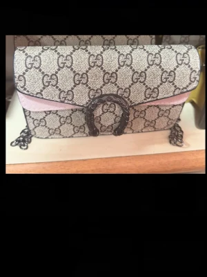  kuvertväska med GG mönster  - Snygg kuvertväska från G u c c i  klassiskt GG-mönster i beige och brunt. Väskan har en rosa detalj på framsidan och en dekorativ metallspänne. Kedjerem i metall och stilren, rektangulär form. Perfekt accessoar för att lyfta din outfit.