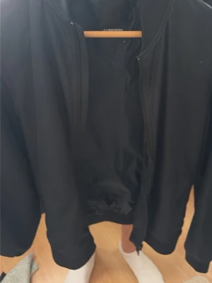 Svart bomberjacka från Jlindberg - Stilren svart bomberjacka från Jlindberg med klassisk passform. Jackan har ribbade muddar vid ärmslut och midja samt dragkedja framtill. Perfekt för dig som vill ha en enkel och snygg jacka till vardagslooken.