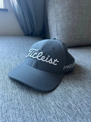 Grå Titleist keps med brodyr - Snygg grå keps från Titleist med broderad logga framtill och på baksidan. Kepsen har böjd skärm, justerbart spänne bak och texten PRO V1 på sidan. Tillverkad i ett lätt och slitstarkt material, perfekt för golf eller streetwear.